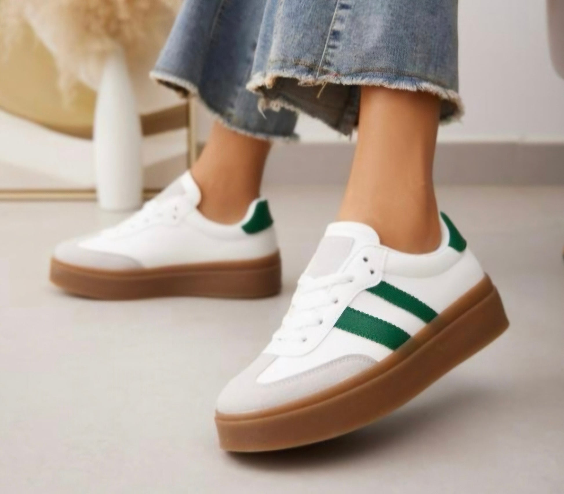 A close-up view of a pair of women's casual trainers. The trainers feature a white body with two green stripes on the sides and a green heel tab. The shoes have a thick, light brown gum sole. The wearer is shown standing on a light-coloured floor, wearing frayed hem jeans.