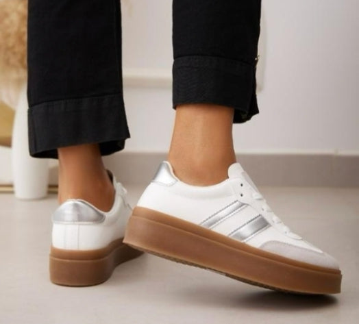 A close-up view of a pair of women's casual trainers. The trainers feature a white body with two metallic silver stripes on the sides and a silver heel tab. They are designed with a thick, light brown gum sole. The wearer is shown standing on a light-coloured floor, wearing black trousers with folded cuffs.